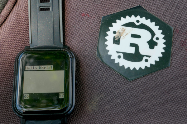 A smart watch showing "Hello World!" and some numbers on a somewhat dirty black textile with a slightly dirty sticker with the Rust logo on the textile